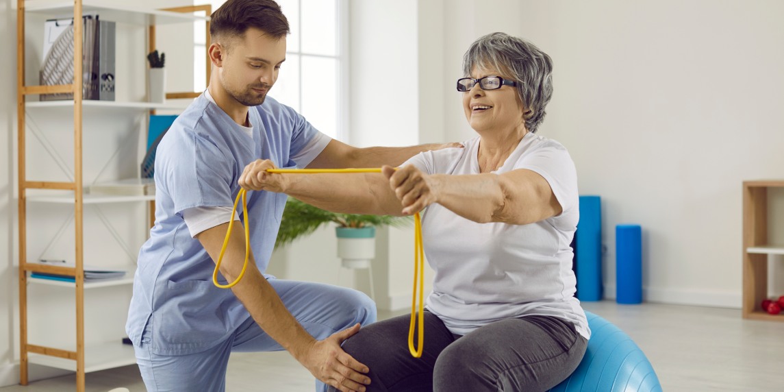 Young doctor at rehabilitation centre helps senior patient do exercises on fit ball. Cheerful old lady with osteoporosis sitting on stability ball, holding rubber band and doing physiotherapy exercise
