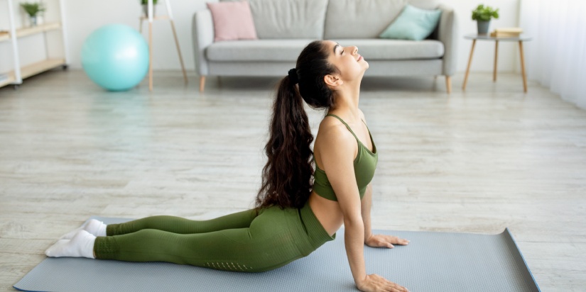 Side view of fit Indian woman doing yoga cobra pose or pilates on mat in living room. Sporty Asian lady working out indoors during covid-19 isolation. Stay home hobbies and active lifestyle concept